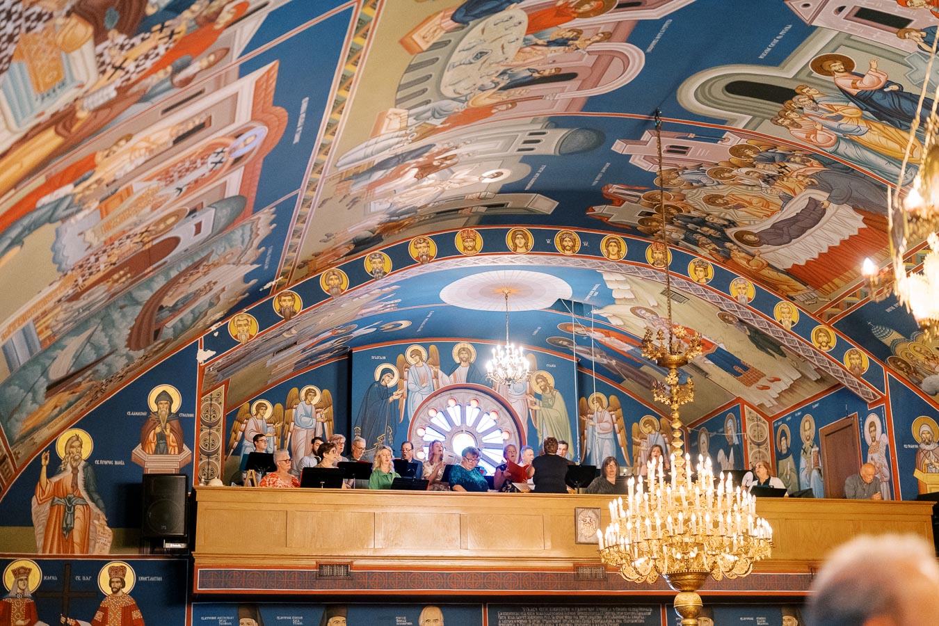 Interior of a church adorned with vibrant religious murals and chandeliers, featuring people in a choir loft, enhancing the spiritual ambiance.