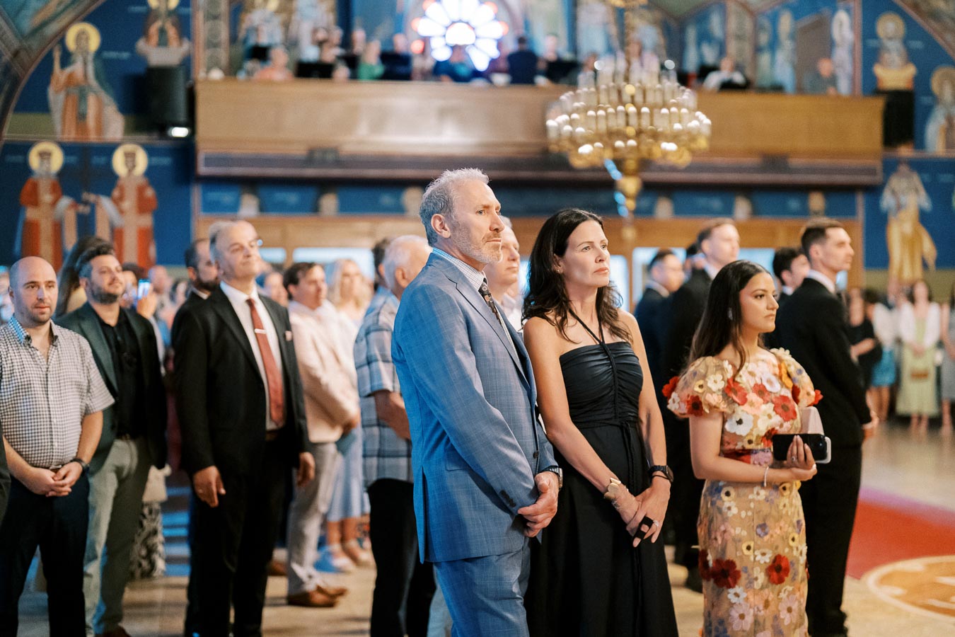 A diverse group of people attentively gathered in a beautifully decorated indoor event space with ornate chandeliers and artistic wall murals.