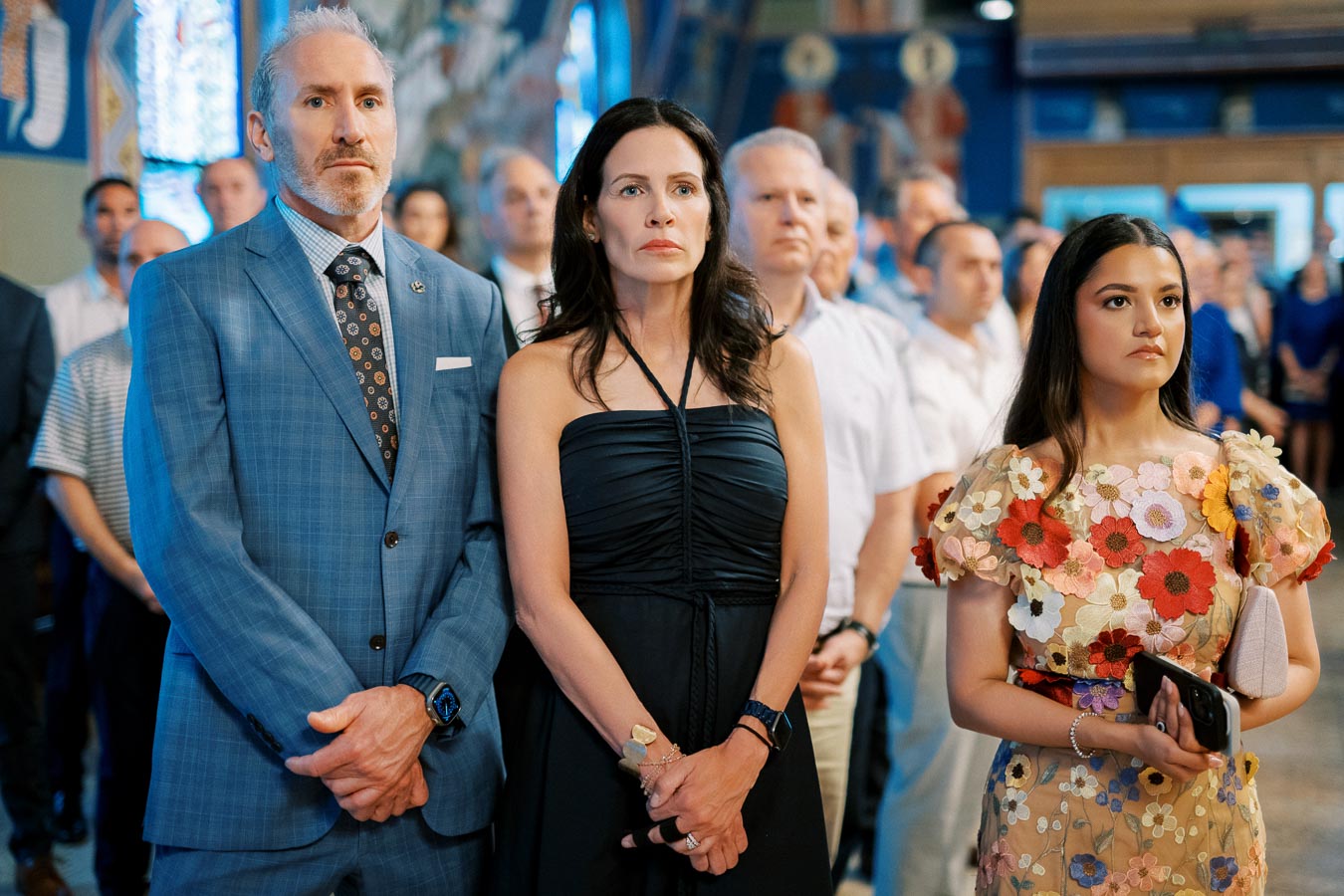A group of people attentively standing indoors, including a man in a blue plaid suit, a woman in a black dress, and another woman wearing a floral-patterned dress, in what appears to be a formal or ceremonial setting.