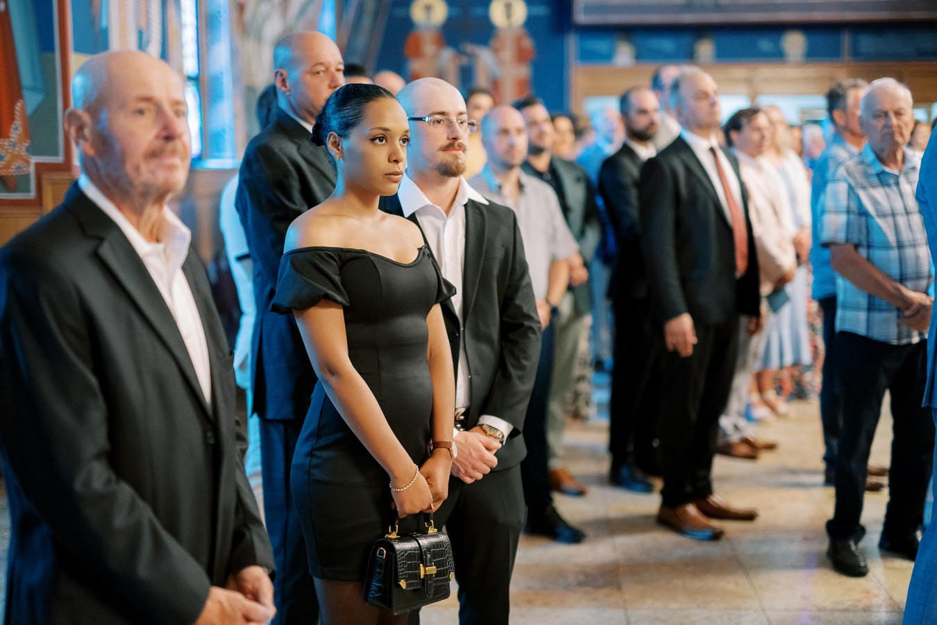 A diverse group of people dressed in formal attire attending a gathering inside a decorated venue, with focus on a woman in a black dress holding a handbag, highlighting a sense of occasion and focus.