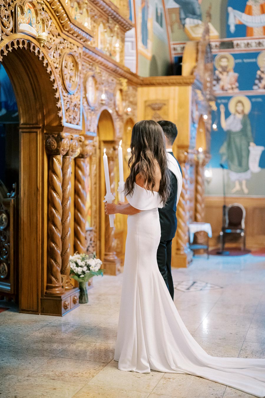 A bride and groom holding candles during a traditional wedding ceremony inside a beautifully decorated church with intricate wooden carvings and religious iconography.