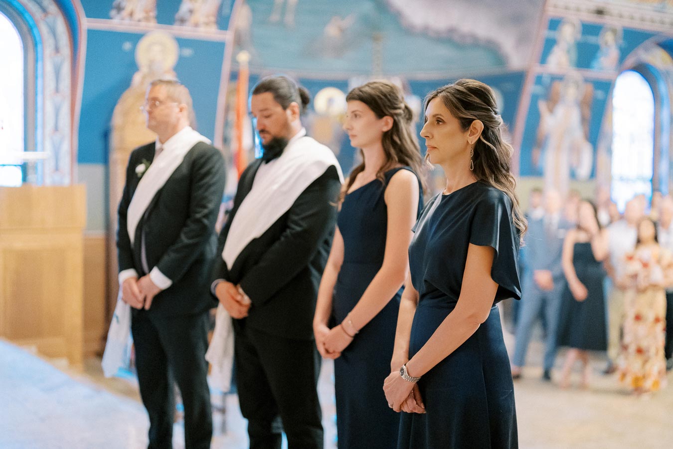 A group of people standing in a church ceremony, wearing formal attire, including navy dresses and suits, framed by a vibrant and ornate interior.