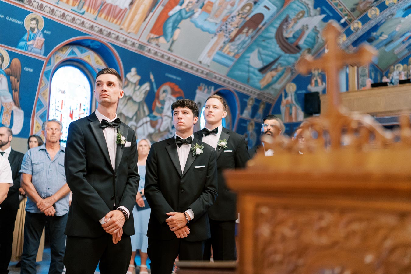 A group of men in black tuxedos with boutonnieres stand inside a colorful, intricately decorated church, captured during a wedding ceremony.