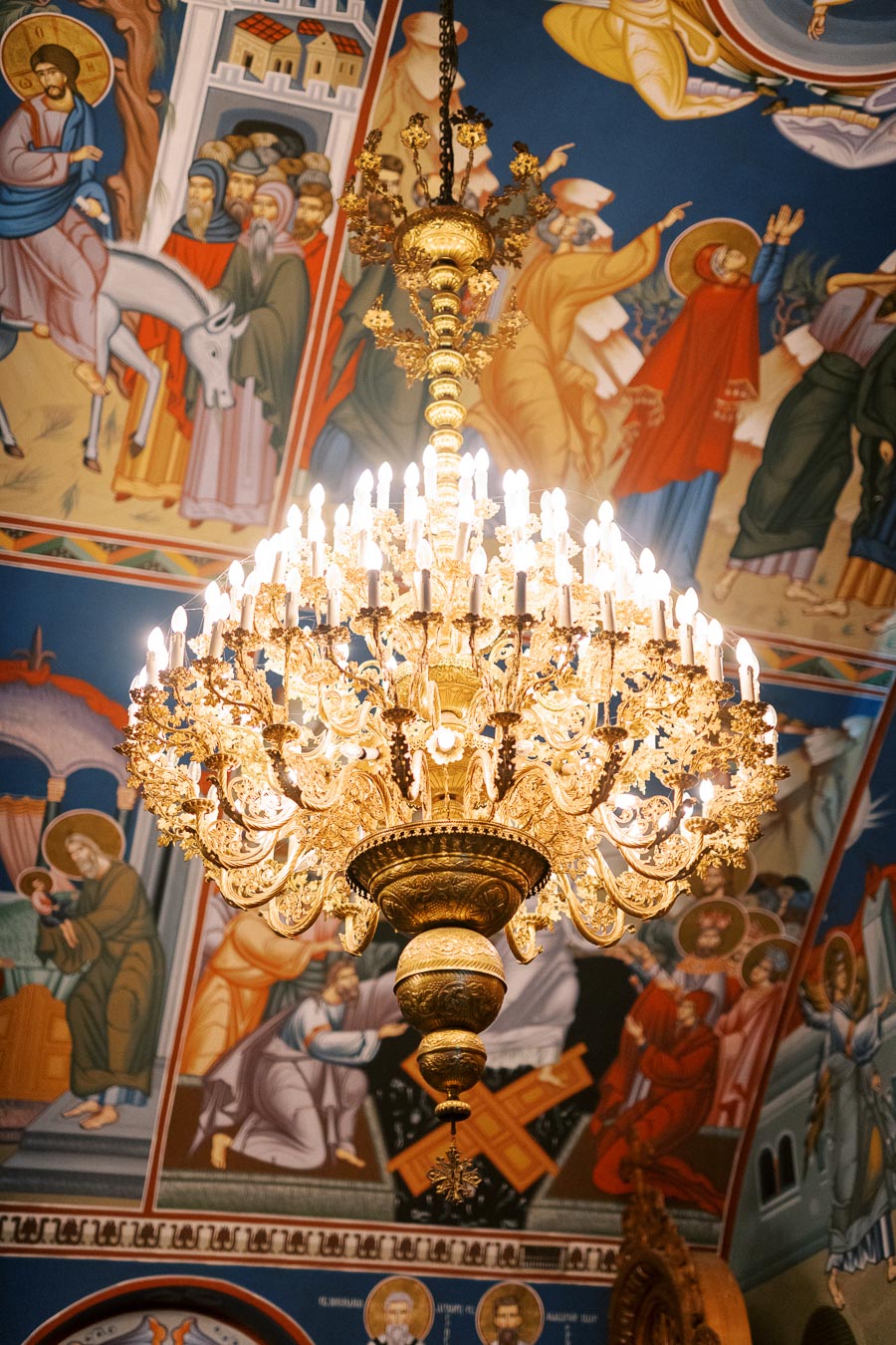 Ornate golden chandelier illuminated inside a church, with vibrant biblical frescoes depicting religious scenes on the ceiling.