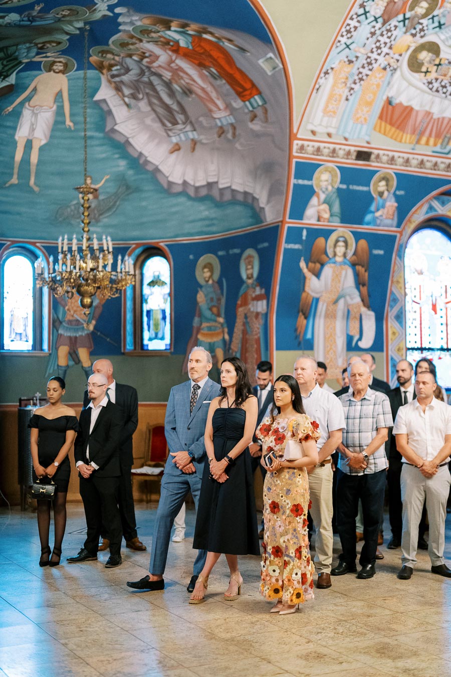 Group of people attending a ceremony in an ornately decorated church, with colorful frescoes and stained glass windows.