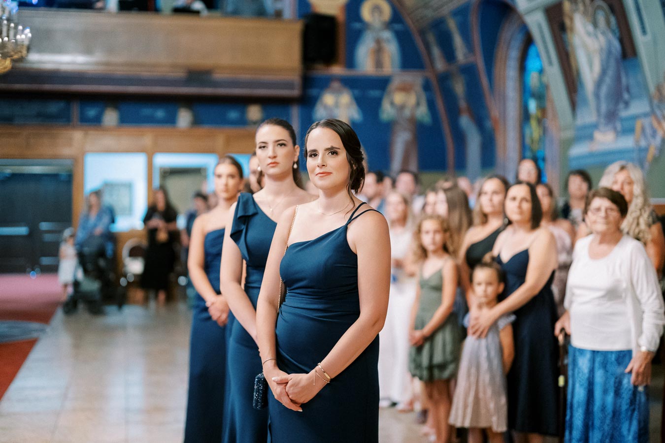 Three bridesmaids in elegant navy dresses stand attentively in a beautifully decorated church, surrounded by a crowd of guests observing a wedding ceremony.