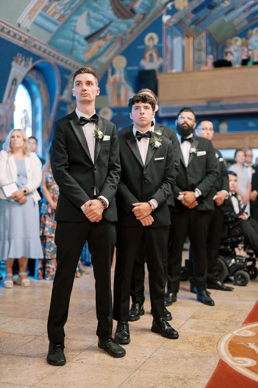 Groomsmen in black tuxedos standing inside a beautifully decorated church with vibrant wall art, attending a wedding ceremony.