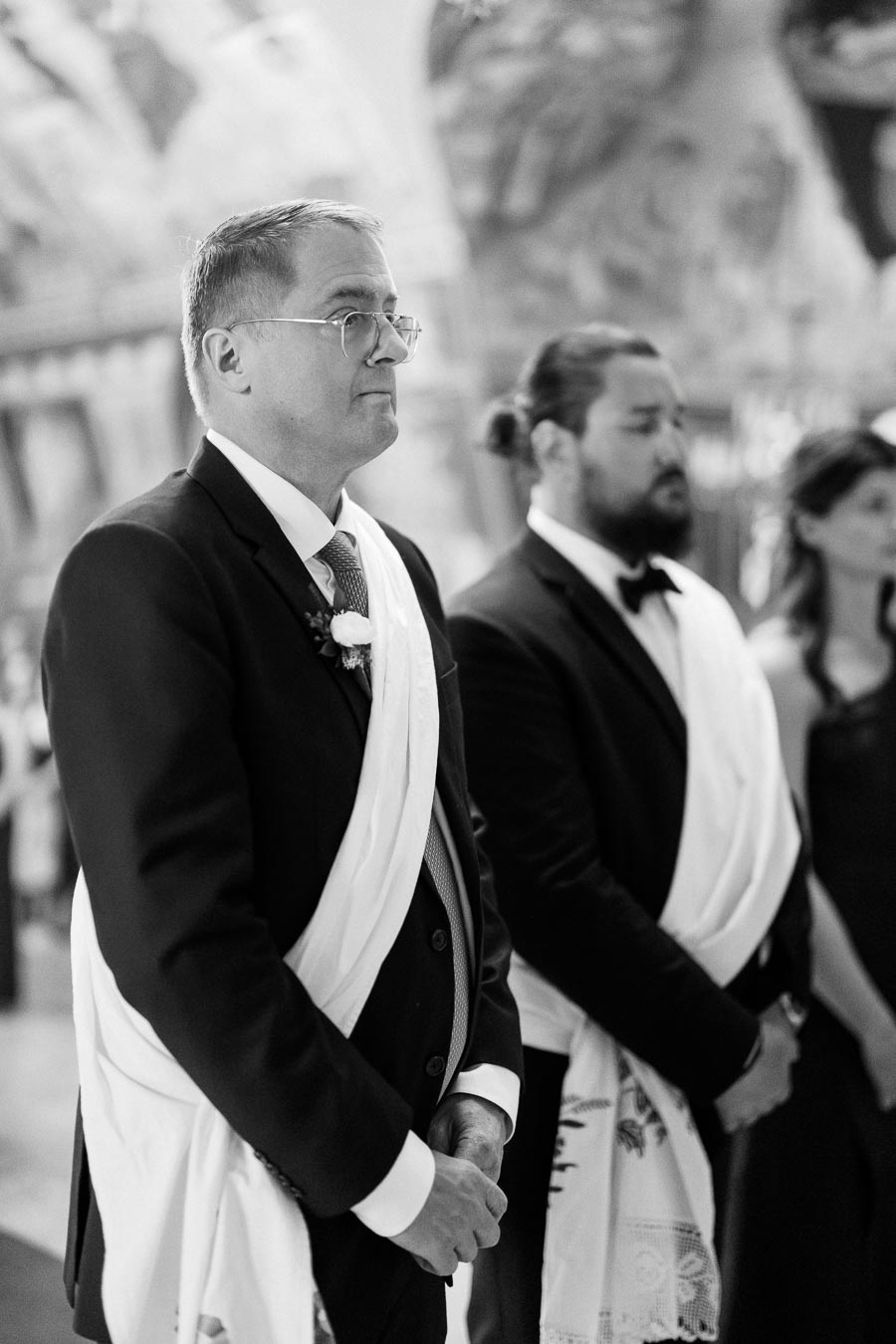 A black and white photograph of two men in suits standing indoors, wearing ceremonial white sashes. The scene suggests a formal event or ceremony with blurred architectural details in the background.