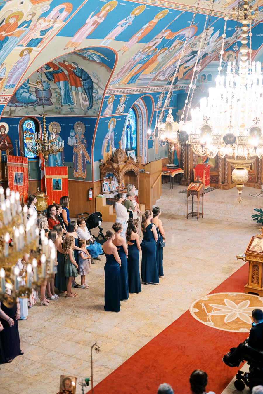 Wedding ceremony inside an ornately decorated church with vibrant frescoes and chandeliers; guests and wedding party dressed formally gather around the altar.