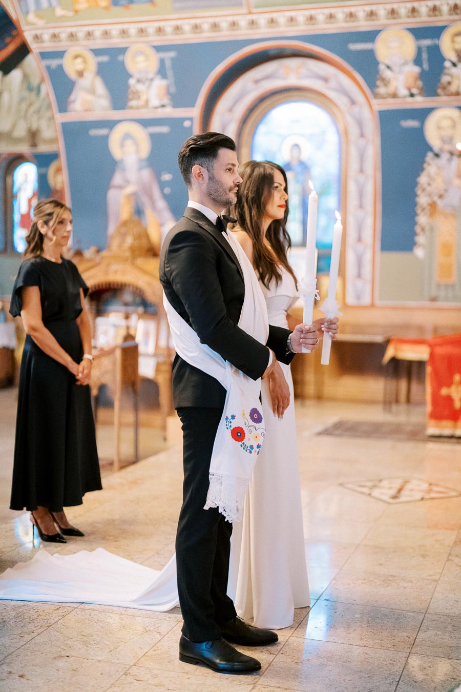 A couple holding candles during a traditional ceremony in an ornate church setting, dressed elegantly in formal attire with embroidered sashes, while a woman stands behind them.