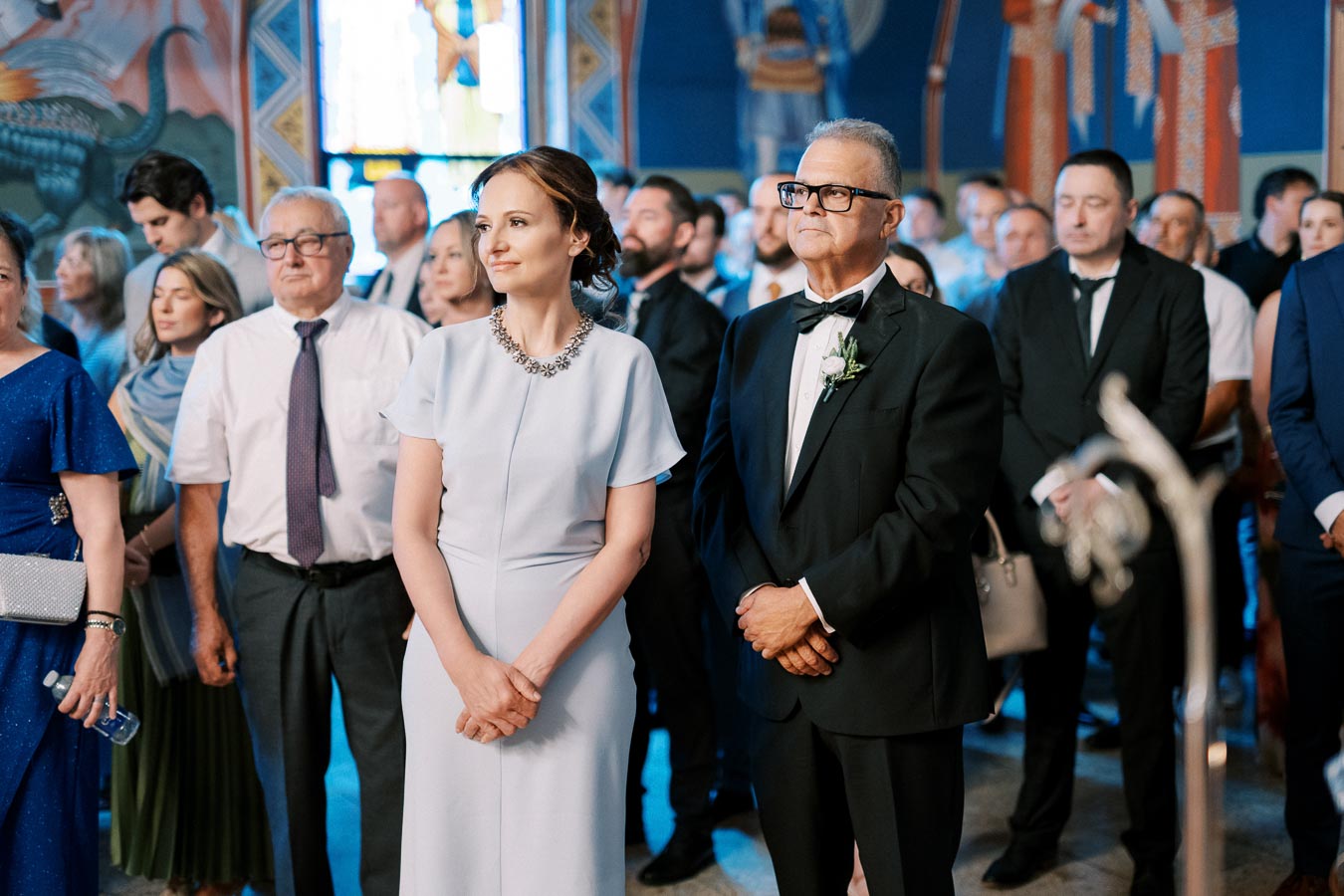 A group of elegantly dressed people attending a formal event inside a beautifully decorated venue with stained glass windows and vibrant wall art.