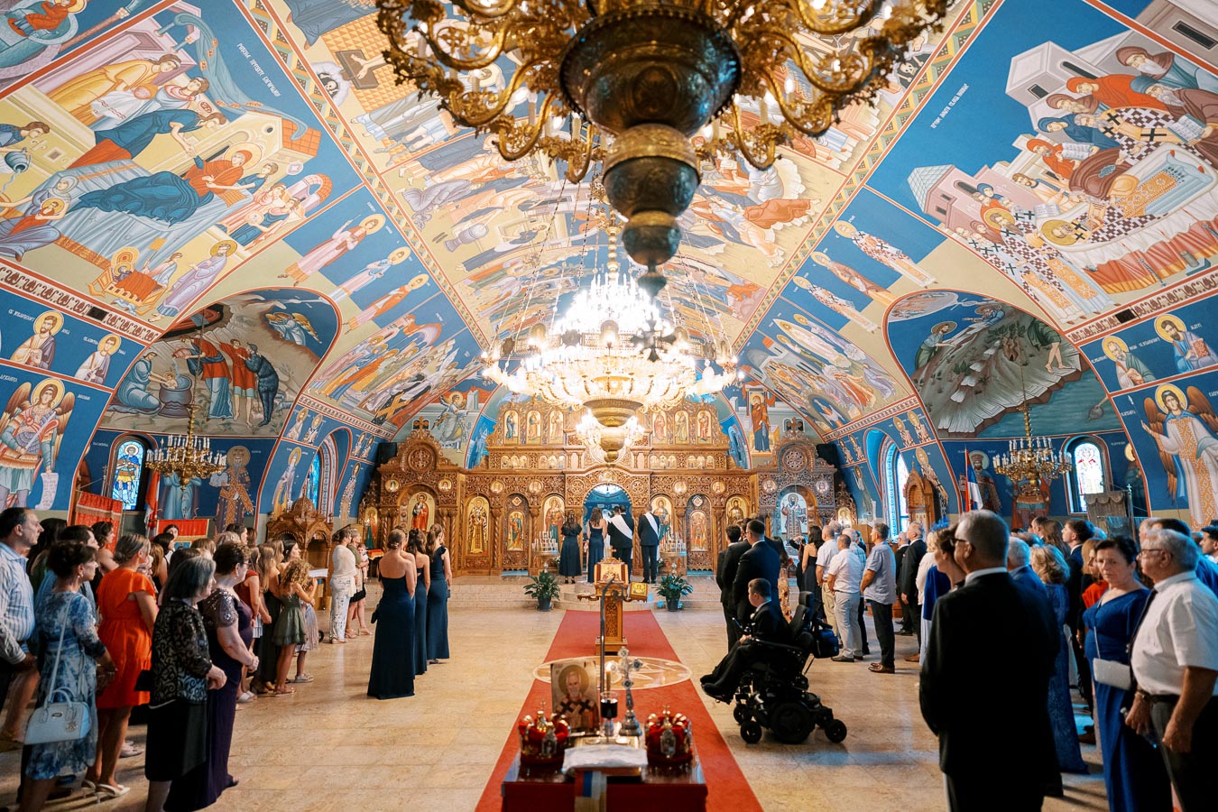 A beautifully decorated Orthodox church interior, featuring vibrant religious frescoes on the walls and ceiling, with chandeliers hanging above. A wedding ceremony is taking place with guests seated in rows, and intricately carved wooden iconostasis visible in the background.