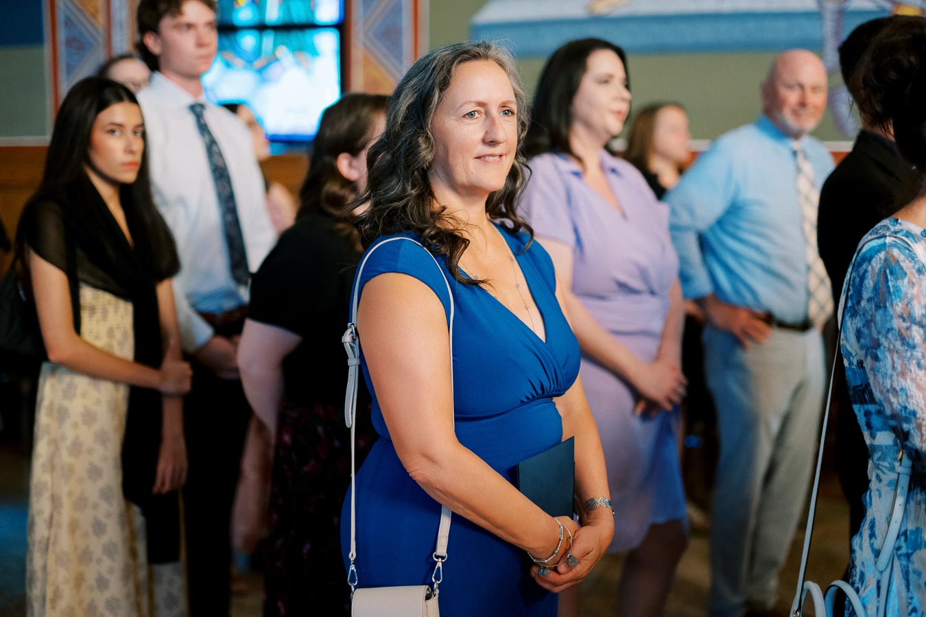 A group of people standing attentively indoors, with a woman in a blue dress in focus, surrounded by others in formal attire.
