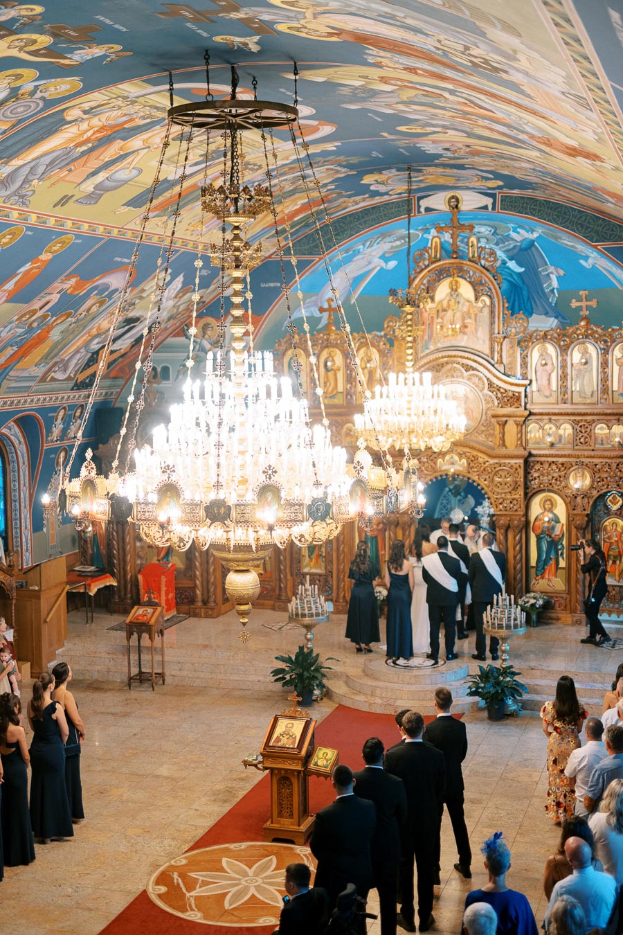 A stunning interior of a beautifully decorated Eastern Orthodox church featuring a grand chandelier, vibrant iconography, and an ornate iconostasis during a wedding ceremony with guests in formal attire.