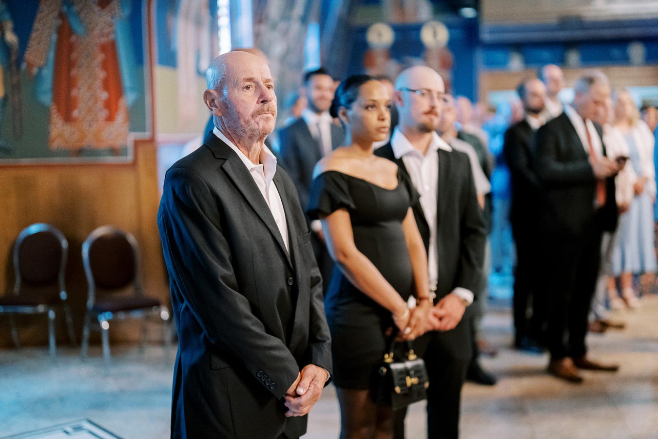 A group of people dressed in formal attire standing attentively in a ceremony setting, with decorated walls and chairs in the background.