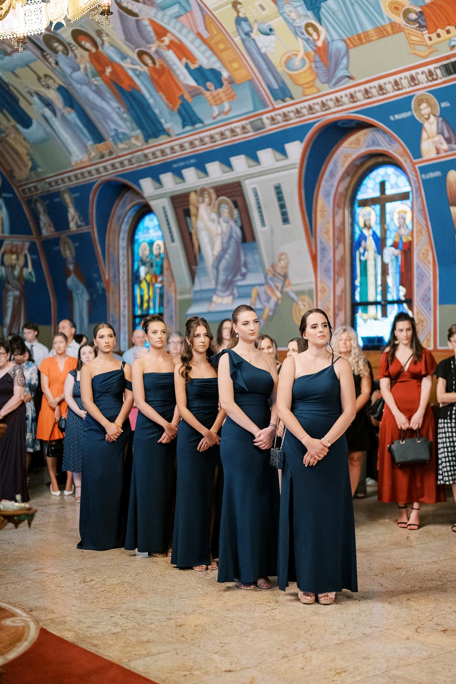 Bridesmaids in matching navy gowns stand in a beautifully decorated church with colorful religious frescoes and stained glass windows in the background, creating an elegant and serene wedding atmosphere.
