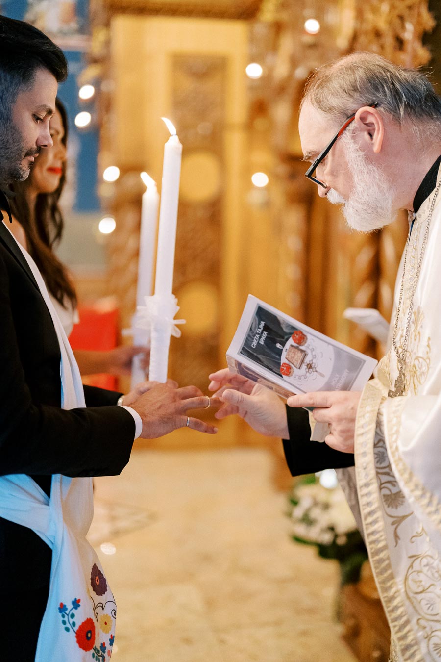 A couple participating in a Greek Orthodox wedding ceremony, exchanging rings in front of a priest who is holding a religious book, with lit candles and ornate decorations in the background.