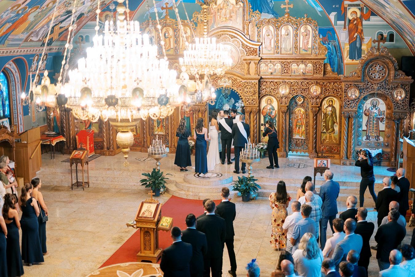 Orthodox wedding ceremony inside a beautifully decorated church with intricate wooden icons and a grand chandelier, guests observing the couple getting married.