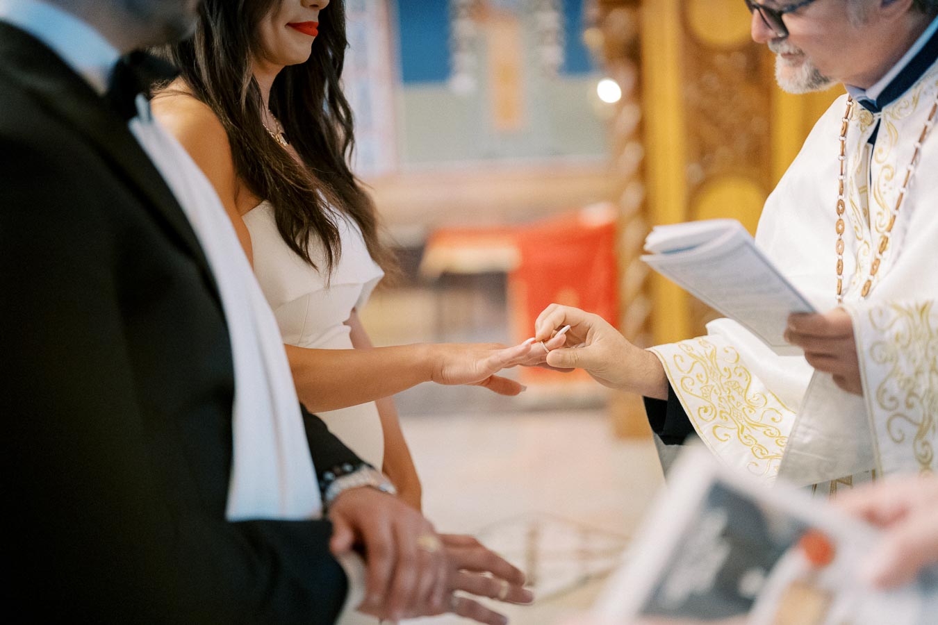 A priest conducting a wedding ceremony as a bride exchanges rings with the groom, with close-up focus on the hands and ceremonial attire.