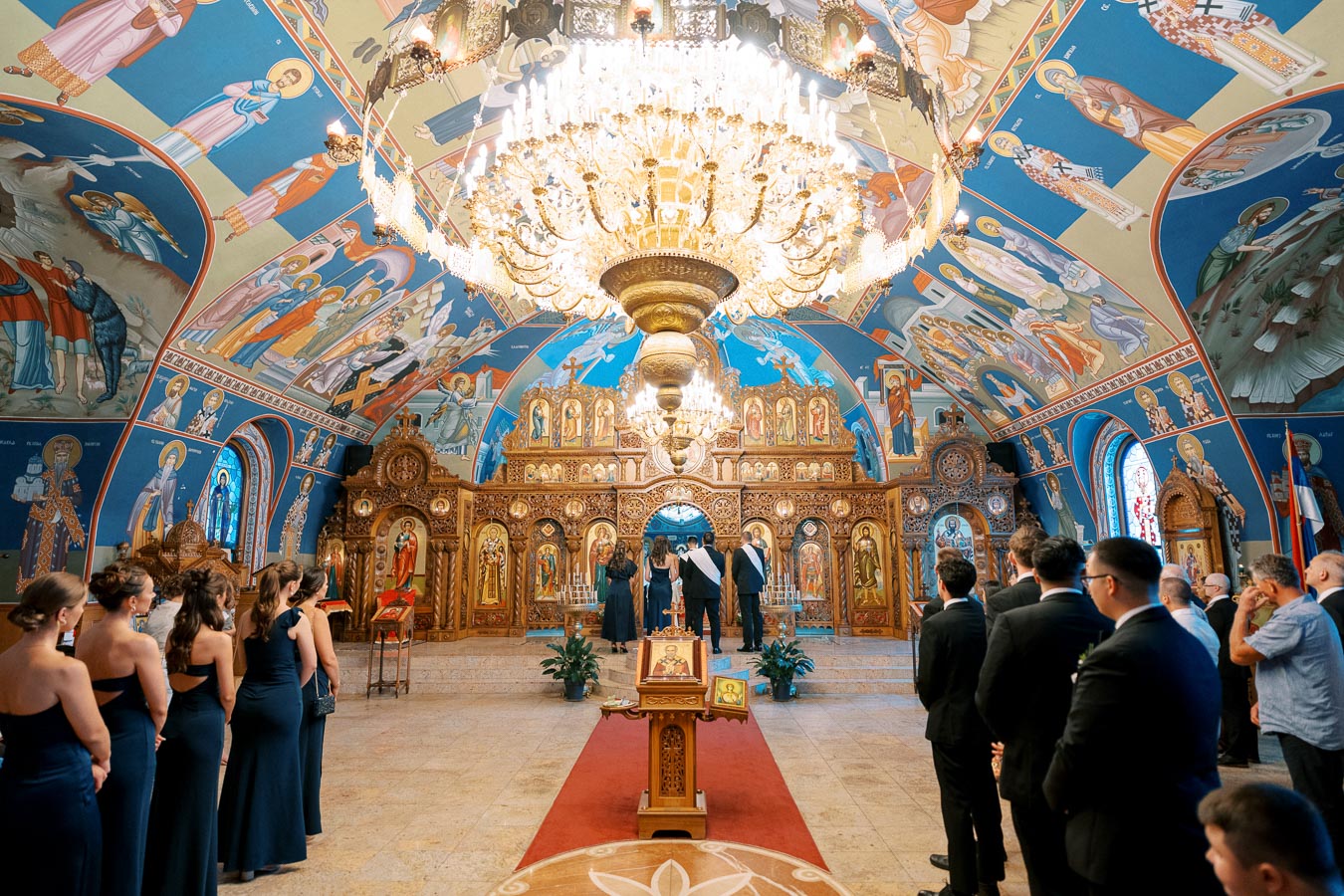 Interior of a beautifully decorated Orthodox church with vibrant religious murals and an ornate chandelier, showcasing a wedding ceremony in progress with attendees in formal attire.