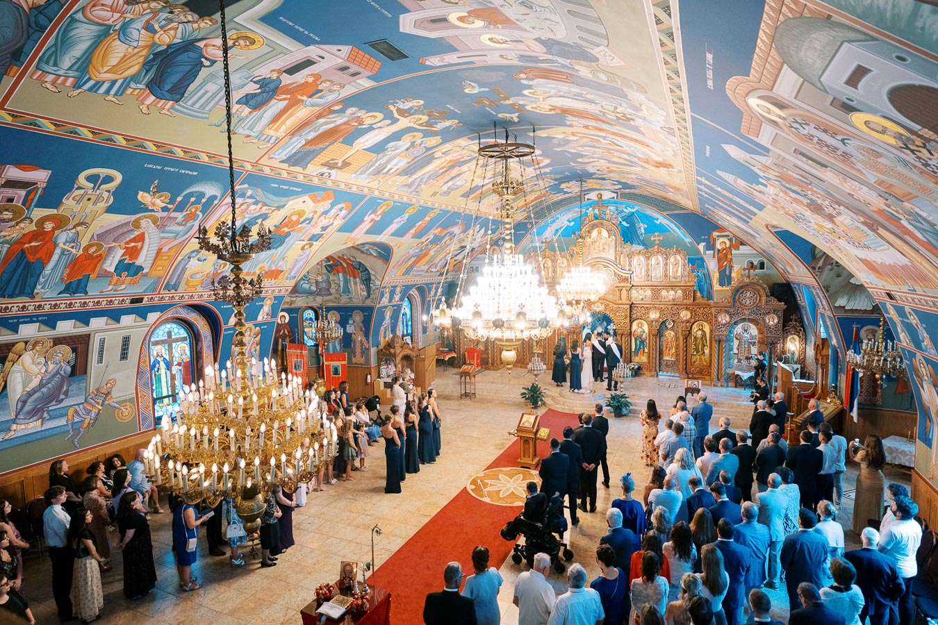 Interior of an ornately decorated Eastern Orthodox church during a wedding ceremony, featuring colorful iconography on the walls and ceiling, a grand chandelier, and an audience of guests gathered around the altar.