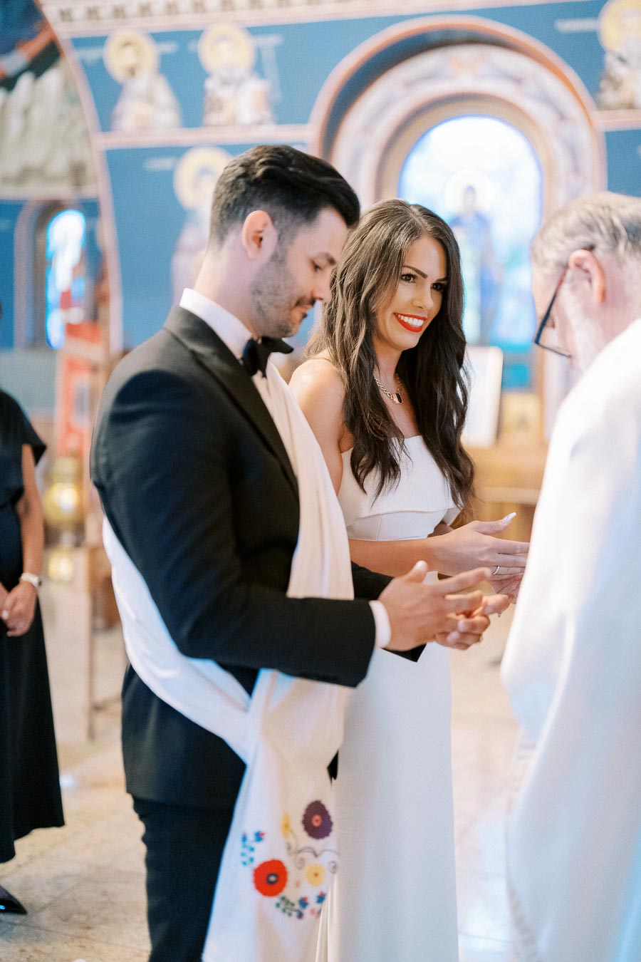 A couple dressed elegantly in a traditional church ceremony, standing before a clergy member. The setting features ornate religious iconography and blue accents, creating a sacred atmosphere.