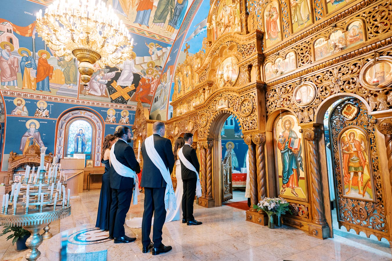 A group of people participate in a traditional ceremony inside a richly decorated Orthodox church, featuring ornate wooden carvings and colorful religious icons. A large chandelier hangs from the ceiling, illuminating the elaborate artwork.