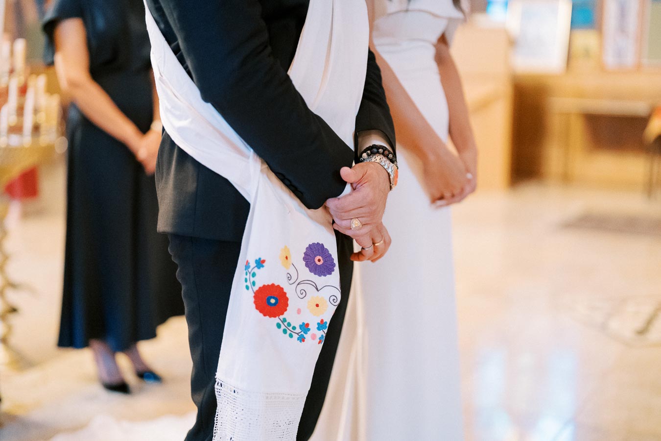 A couple stands in a ceremony wearing traditional attire; a man in a black suit holds a white embroidered scarf with colorful flowers, while a woman in a white dress stands beside him, highlighting a cultural or religious event setting.