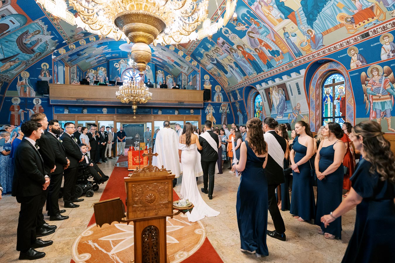 A wedding ceremony inside a beautifully decorated Greek Orthodox church, featuring vibrant wall murals and a large chandelier. The bride and groom stand before the altar, surrounded by groomsmen and bridesmaids in formal attire, as guests watch in the background.