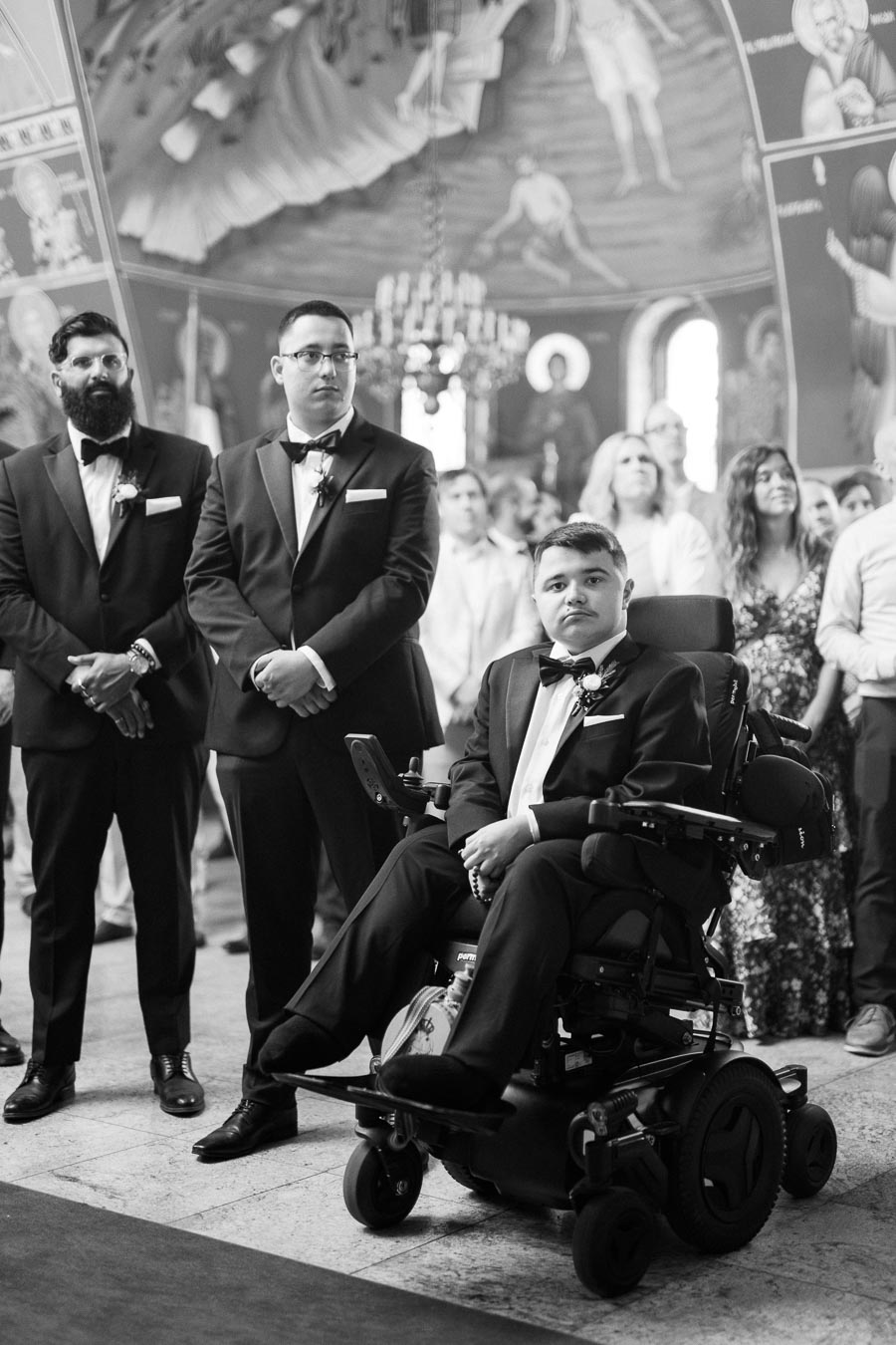 A groom in a wheelchair accompanied by groomsmen in black suits at a wedding ceremony inside an ornate church, surrounded by guests.