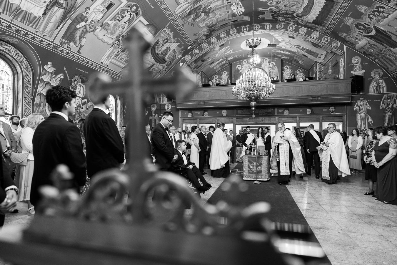 Black and white image of a traditional religious ceremony inside an elaborately decorated church with ornate ceiling artwork. Priests in robes stand near the altar, surrounded by attendees in formal attire, highlighting the solemnity and reverence of the event.