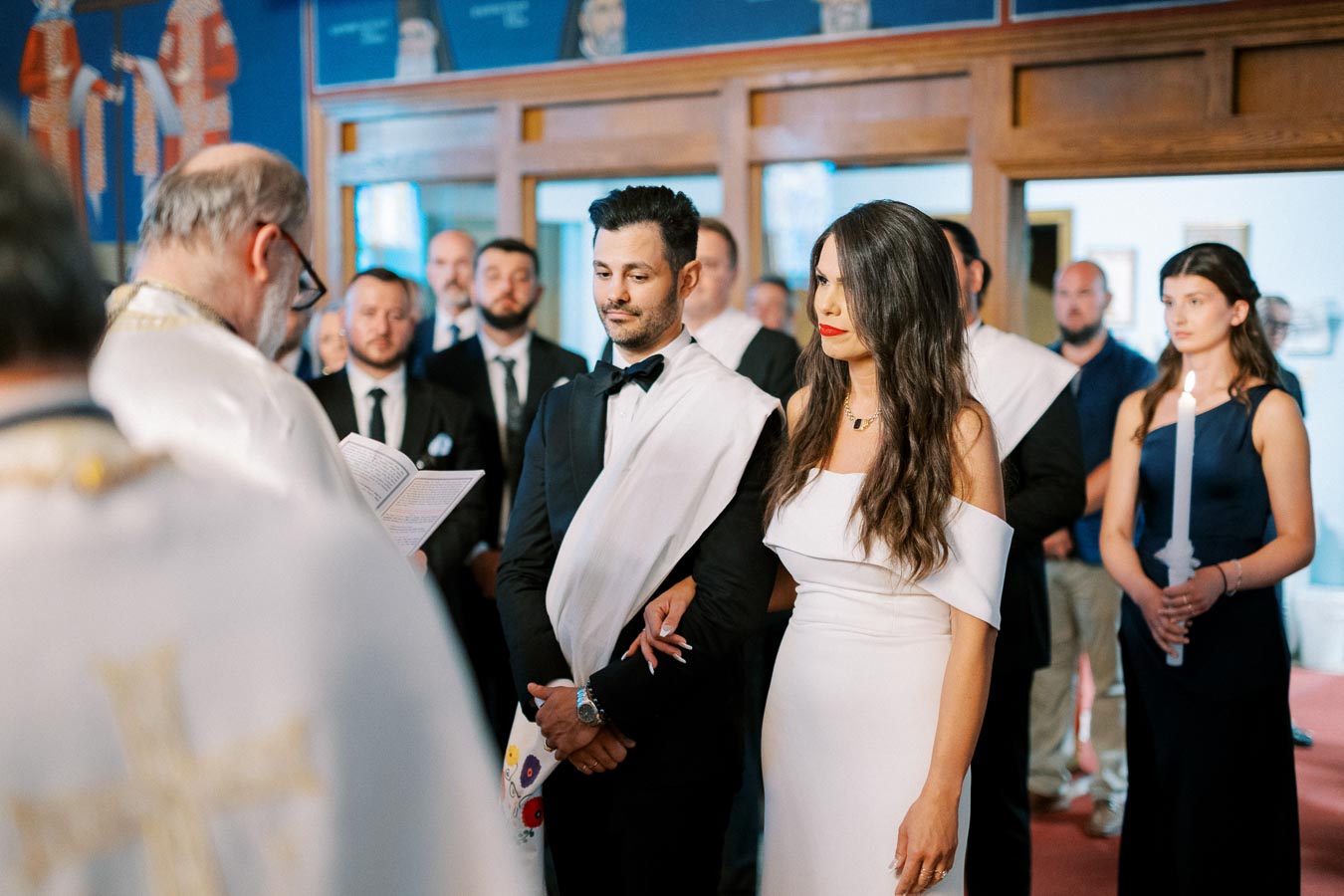 A couple stands together at a wedding ceremony in a church, with a priest officiating the service. The man is in a black suit with a white stole, and the woman is in a white dress. The room is filled with guests and there is a blue wall in the background adorned with images.