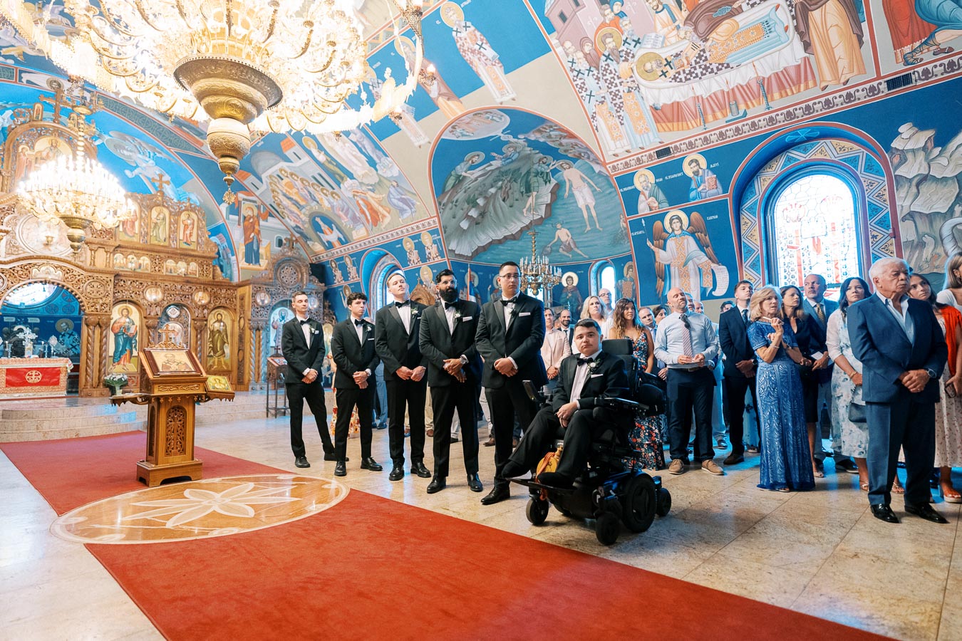 A vibrant wedding ceremony in a beautifully decorated Orthodox church with ornate murals and chandeliers, featuring a group of groomsmen in black tuxedos standing beside a person in a wheelchair.