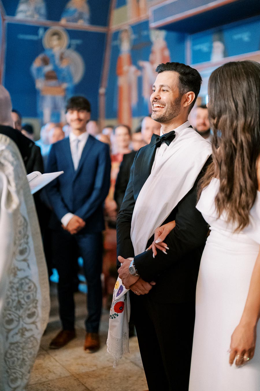Wedding ceremony in a beautifully decorated church, featuring a smiling groom in a tuxedo and a bride in a white dress, surrounded by attentive guests and vibrant murals.