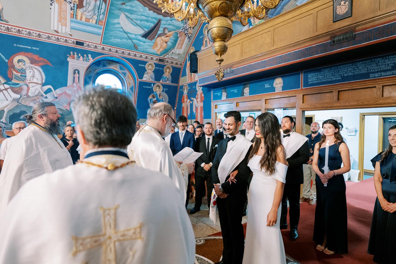 A Greek Orthodox wedding ceremony in a beautifully decorated church, featuring a bride and groom standing before a priest, surrounded by guests. The ornate interior includes vibrant religious paintings and icons on the walls.