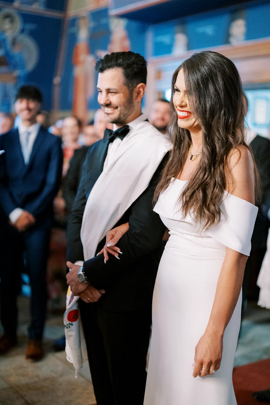 Woman in elegant white dress and man in black suit with a white shawl, smiling during an indoor ceremony with a mural in the background.
