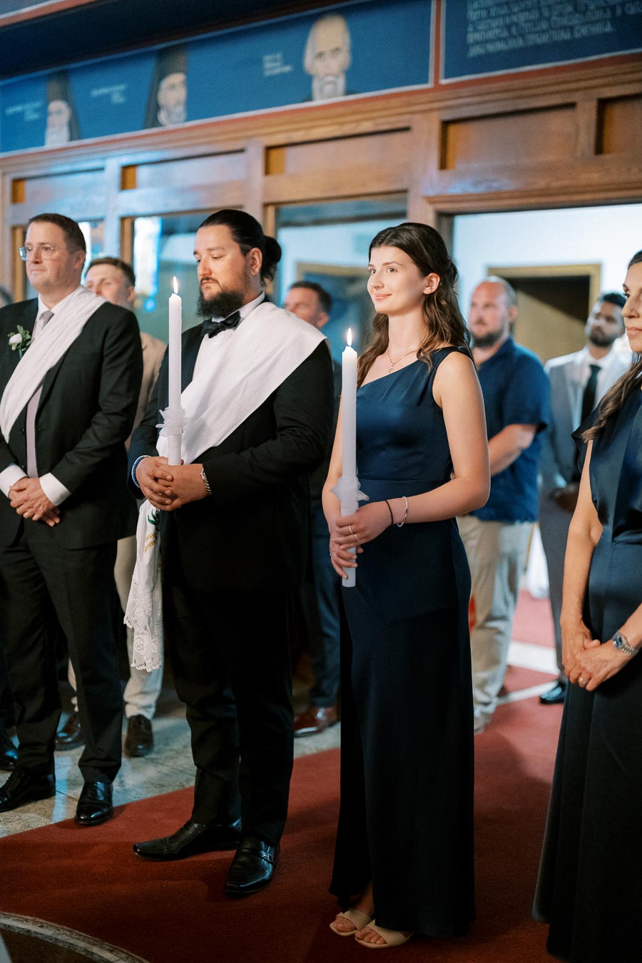 A group of elegantly dressed people standing indoors, holding white candles during a formal ceremony, with wooden interior details and portraits in the background.