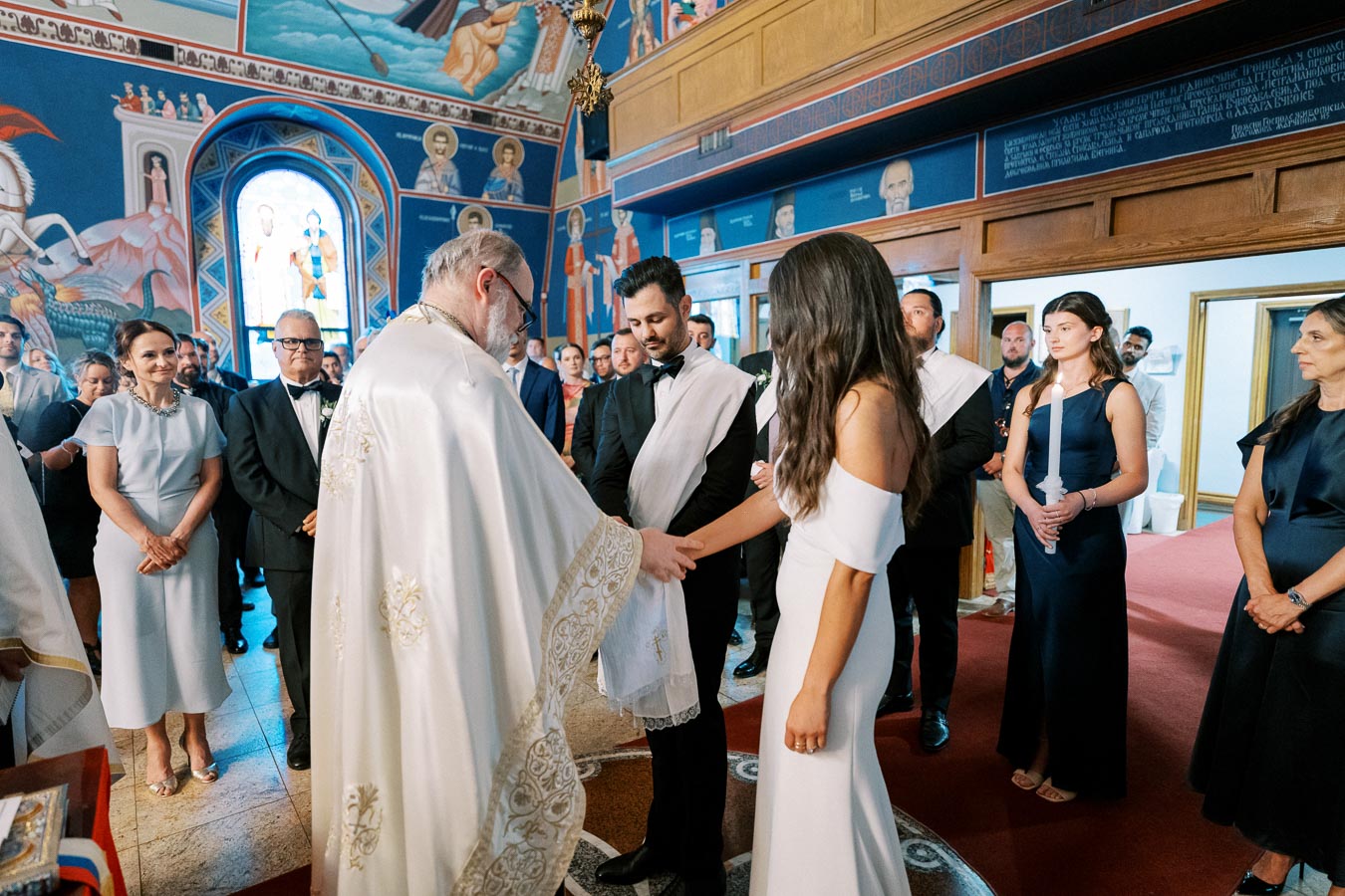 Orthodox wedding ceremony inside a beautifully decorated church, with a priest blessing the bride and groom as guests observe. The bride wears an elegant off-the-shoulder gown, and the groom is in a classic suit, both surrounded by family and friends in formal attire.