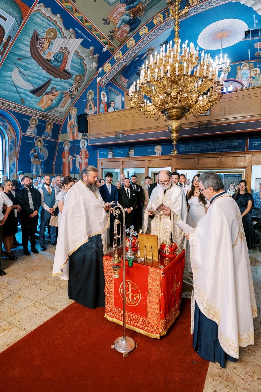 Ceremony inside a beautifully decorated Orthodox church, featuring priests in traditional garments, vibrant religious murals on the walls, and a large golden chandelier above.