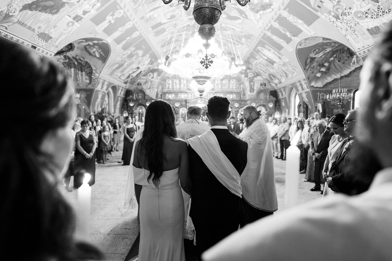 A couple standing at the altar during a traditional wedding ceremony in an ornately decorated church, surrounded by family and friends, captured in black and white.