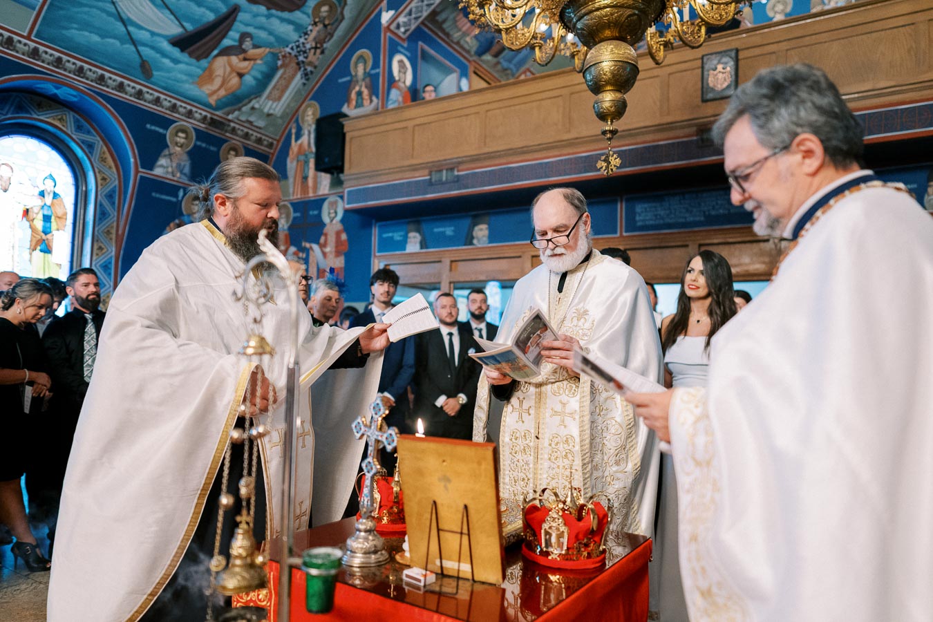 Ceremony in an ornate Orthodox church with priests in white vestments, surrounded by attendees. Iconography and religious artifacts create a traditional atmosphere.