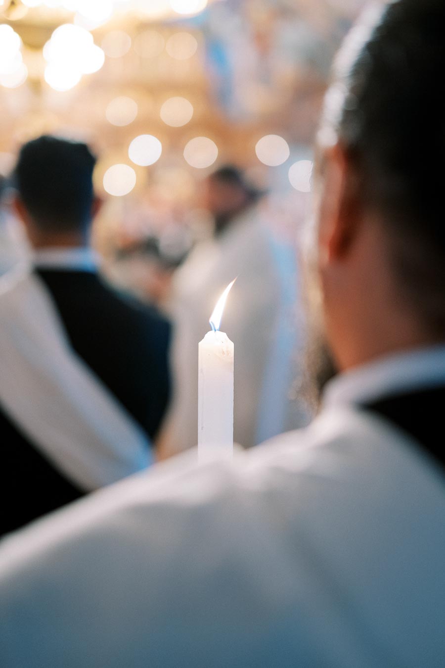 Close-up of a lit candle held by a person during a religious ceremony, with blurred figures in the background, creating a serene and spiritual atmosphere.