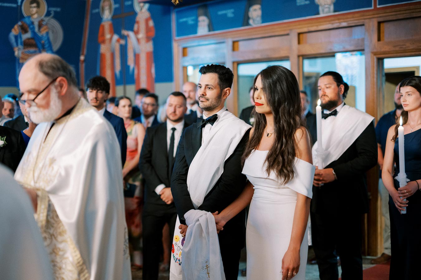 A couple standing inside a church, participating in a wedding ceremony. The man and woman are dressed elegantly in formal attire, holding a white cloth together. The background includes guests and religious artwork on the walls, creating a traditional and solemn atmosphere.