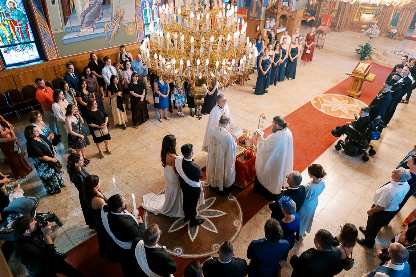 A traditional wedding ceremony inside an ornate church, with a bride and groom standing at the altar surrounded by guests and clergy, under a large chandelier, with stained glass windows and religious iconography in the background.