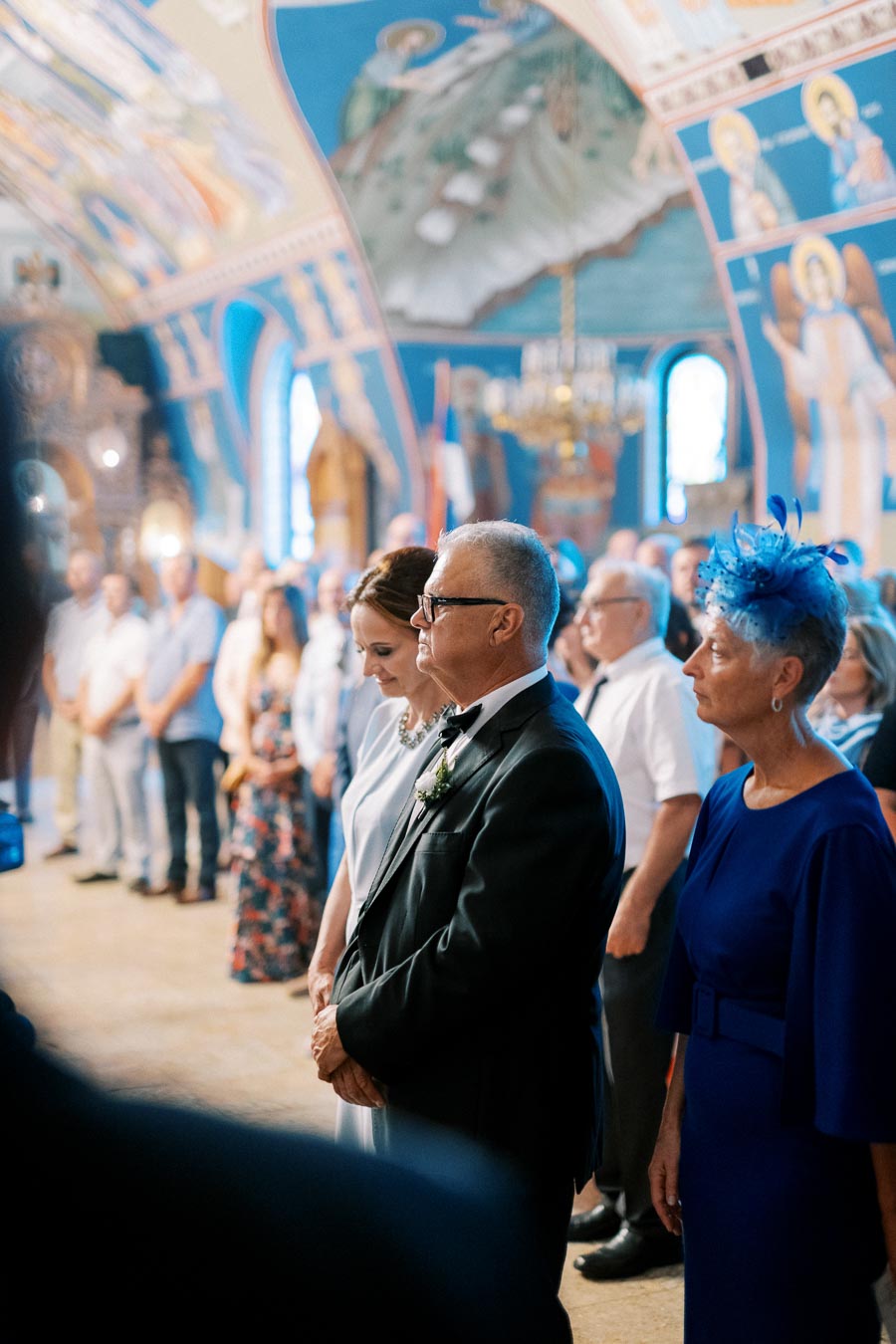 A wedding ceremony inside a beautifully decorated church with attendees in formal attire, featuring ornate murals on the walls and a chandelier in the background.