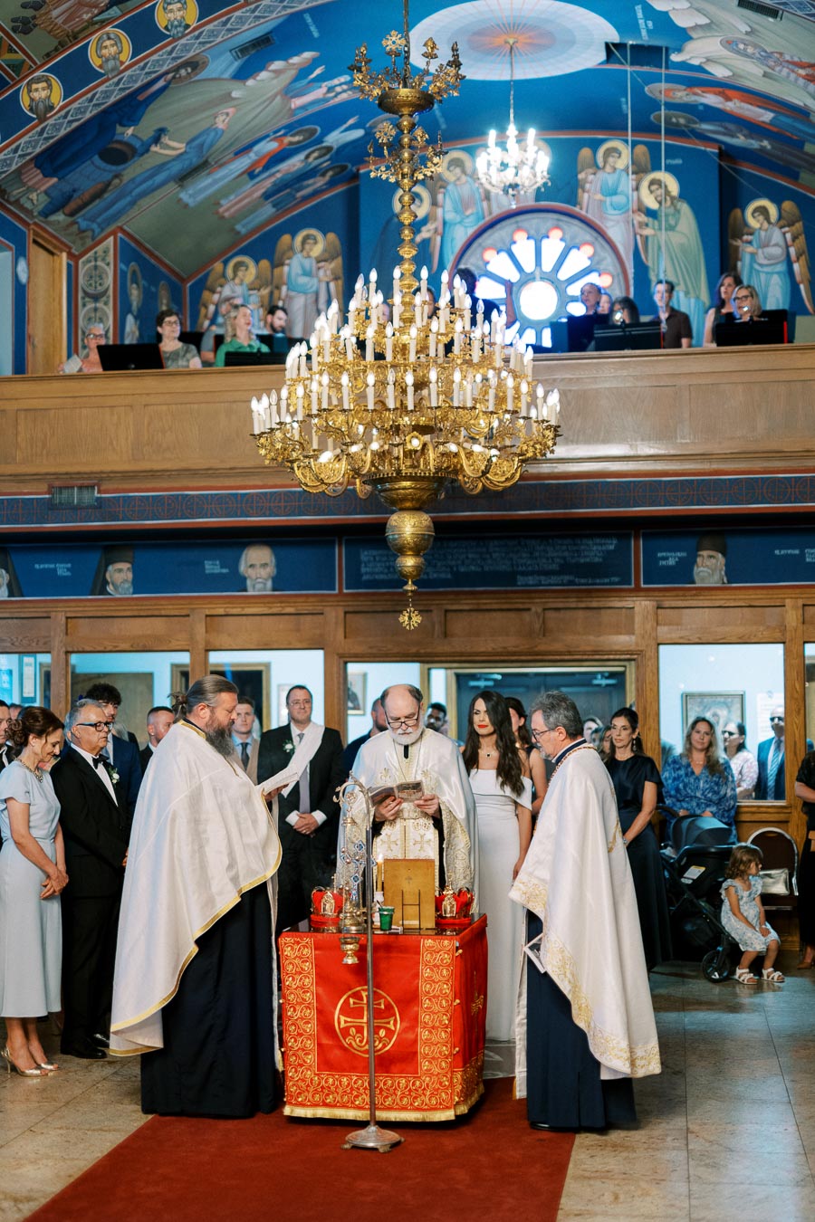 Greek Orthodox wedding ceremony inside a decorated church, featuring a large chandelier, priests in traditional robes, and a gathering of attendees.