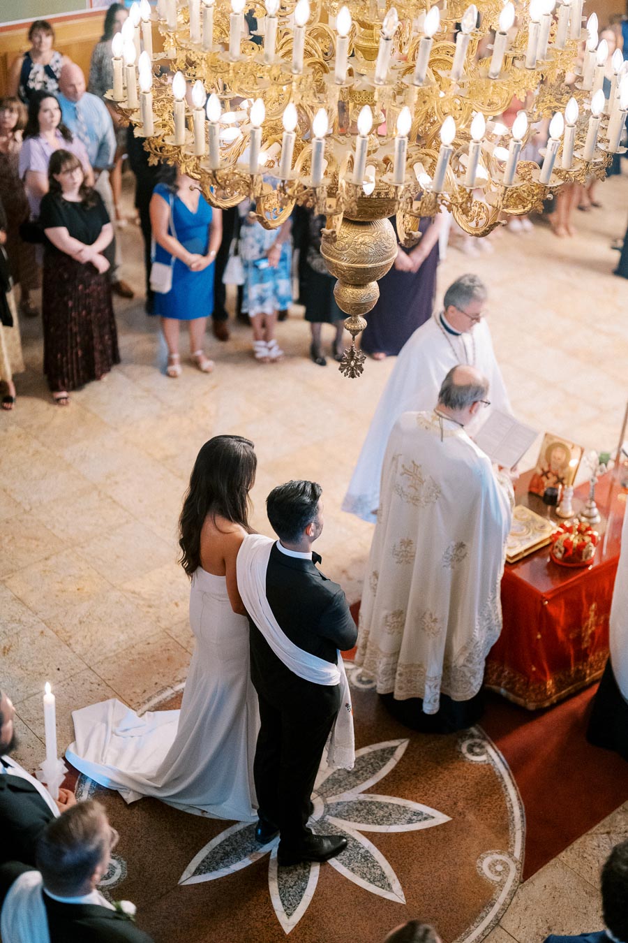 Overhead view of a wedding ceremony in a church, featuring a couple standing together near the altar with a priest, surrounded by guests and illuminated by a large ornate chandelier.