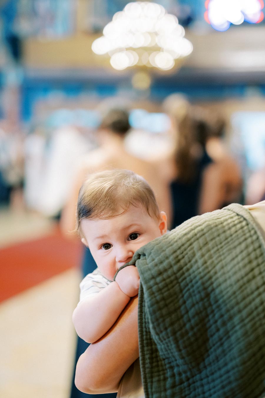 A baby gazes curiously while clutching a green blanket, surrounded by a softly blurred background and warm ambient lighting indoors.