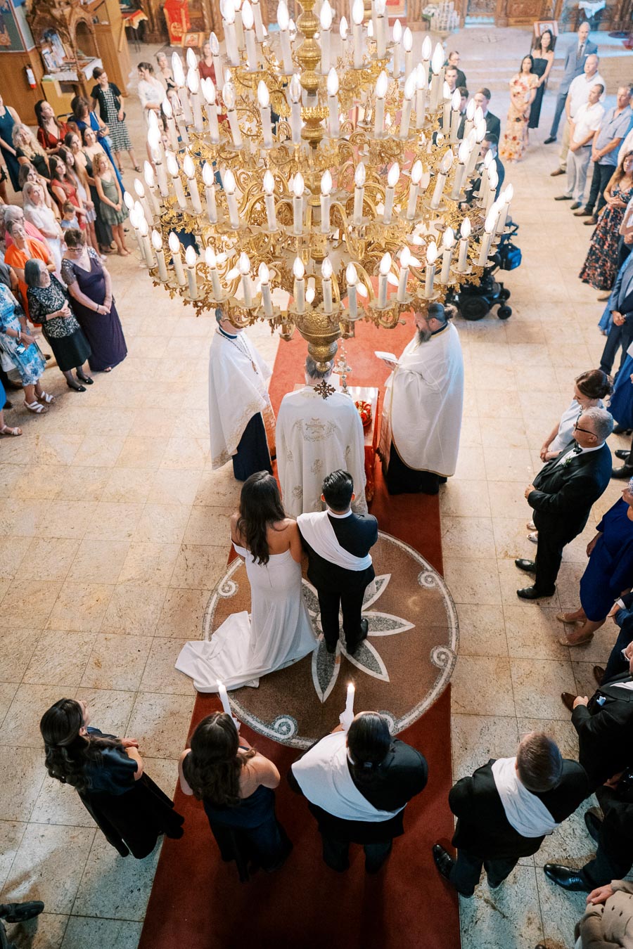Aerial view of a traditional wedding ceremony inside an ornate church, featuring a bride and groom in white attire standing under a large, golden chandelier. Guests surround the couple, observing the ceremony, as two clergy in white robes officiate under the warm glow of the candles.