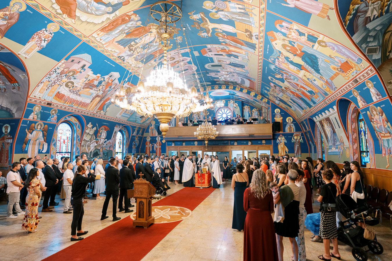 A large group of people gather inside an ornately decorated Orthodox church with vibrant religious frescoes on the walls and a grand chandelier hanging from the ceiling, attending a ceremony or event.